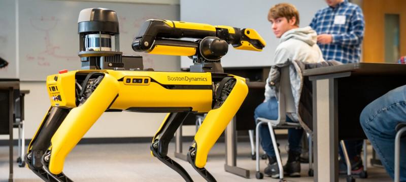 Student in the background at a desk turned around and looking at the yellow and black robot dog.