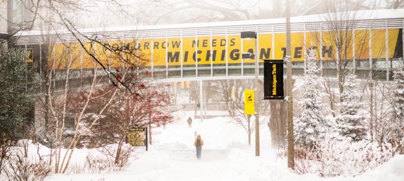 In winter, snow covers the ground under the raised, enclosed walkway between Van Pelt and Opie Library and Rekhi Hall, with signage reading â€œTomorrow Needs Michigan Tech.â€