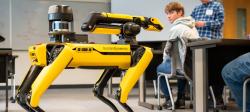 Student in the background at a desk turned around and looking at the yellow and black robot dog.