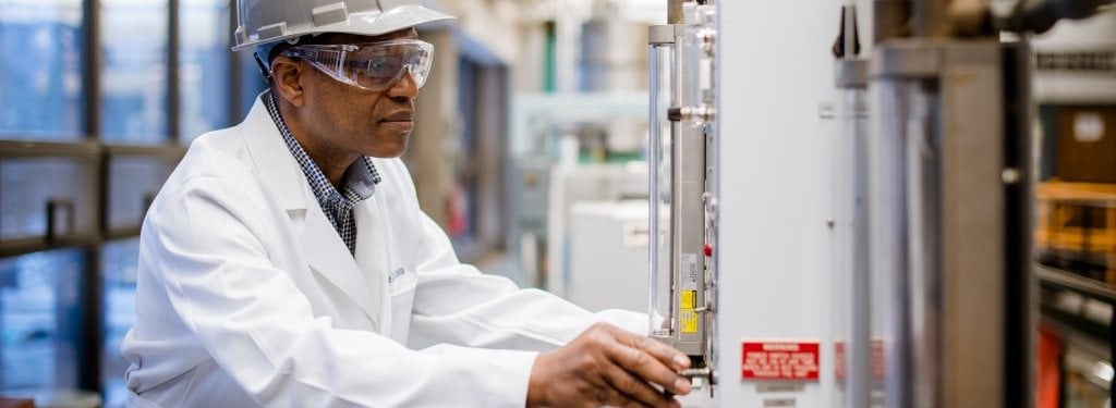 Andre Da Costa in the lab. A man, Andre Da Costa, wearing safety googles, a hard hat, and a white lab coat turns a knob on a machine in a research lab.