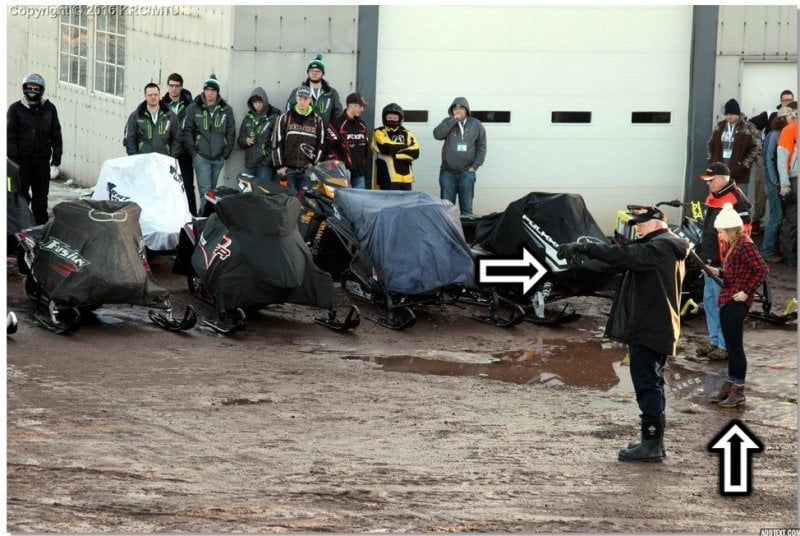 A man in black boots and a young woman in flannel with a winter hat stand in front of a line of people and snowmobiles; two arrows point to them.