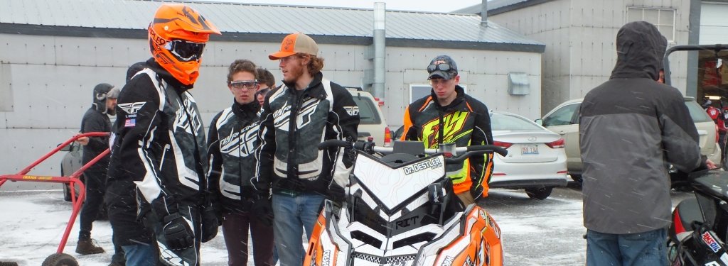 Talking to engineers and snowmobilers is a mix of science and art. A group of engineering students stand around an orange snowmobile.