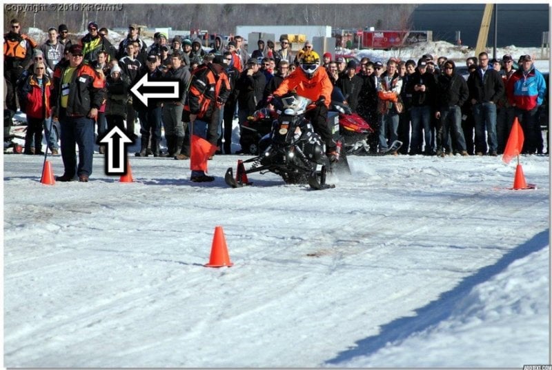 A snowmobile races down an obstacle course and two arrows point to a young woman in the crowd.