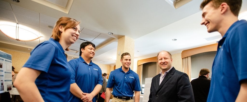 The Medtronic multi-coil passive recharging prototype device Senior Design team chats with sponsor rep and fellow Husky, Carl Wahlstrand, engineering program director for patient care devices with the restorative therapies group. A young woman and three young men all in blue shirts talk with a businessman in a suit in a ballroom with a poster in the background