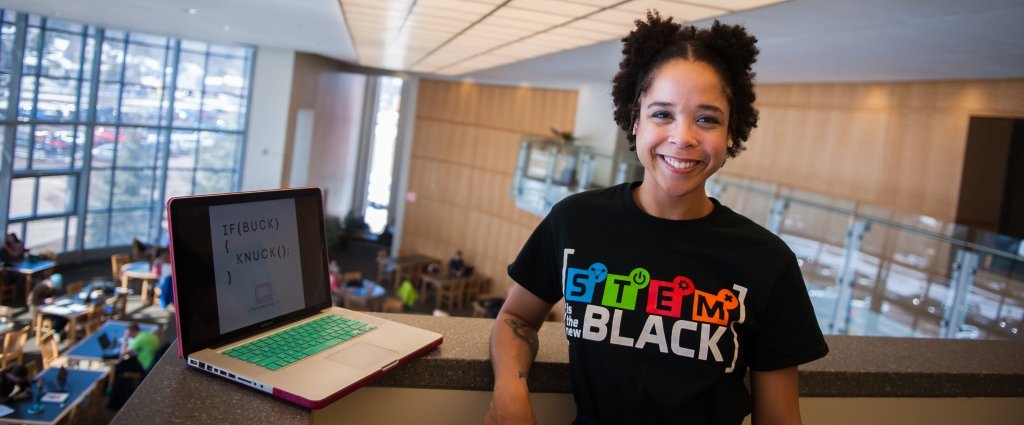 Computer Science PhD candidate Jamie Berger makes code accessible in a new way. On her screen, 2004 hip hop/crunk, "Knuck if You Buck," a post from her Lyrics 2 Code social media project. a young woman with a computer in a library on a floor overlooking another floor where students are studying at tables.
