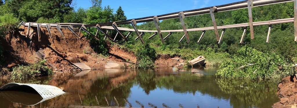 The bridge at Paradise Road over the Pilgrim River washed out during the Father's Day Flood. Several USGS river gauges were swept away in the flooding. Image Credit: James R. Melchiori Washed-out bridge supports hang suspended over a river. A washed-out culvert floats in the water.