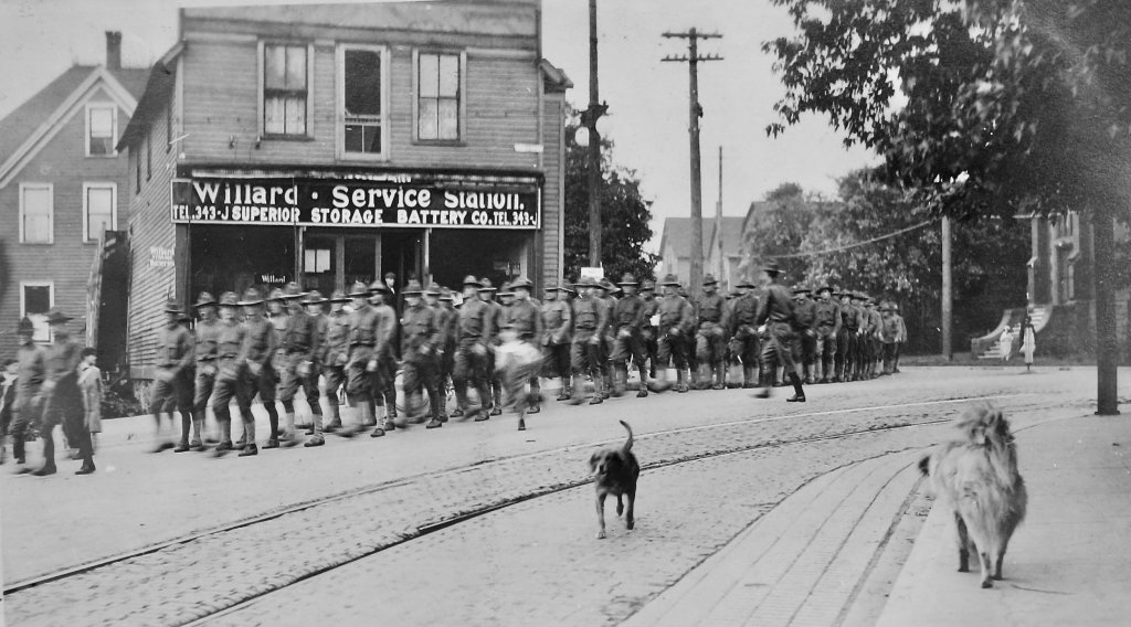 The first detachment to complete mining and military training at Michigan Tech turns onto Shelden Avenue for their final march through downtown Houghton. (National Archives photo) 200 World War I era soldiers march down a cobbled street in 1918 with two dogs in the foreground, one on a sidewalk, and a sign on a storefront in the background that says Willard Service Station.