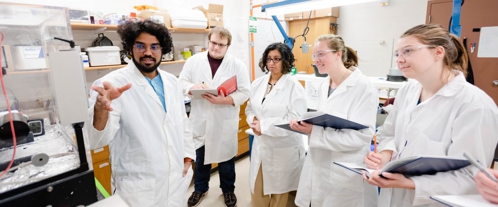 Teamwork makes the dream work — and engineering, too. group of people standing in lab wearing coats and protective glasses