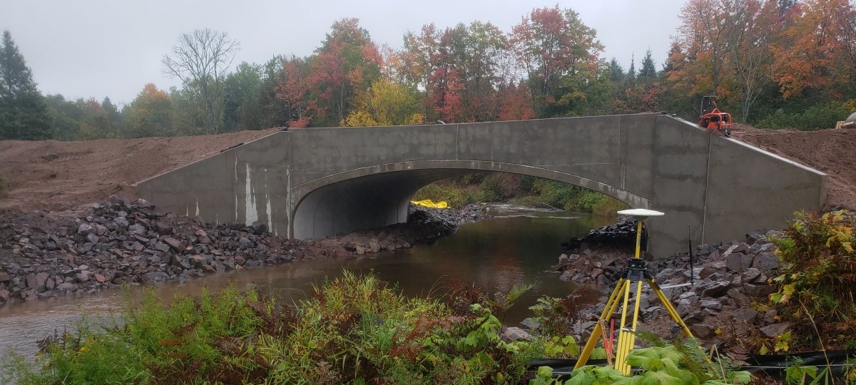 A stream gauge will be placed in spring 2020 by the newly replaced Paradise Road bridge over the Pilgrim River. A bridge crosses a river. The ground around it is bare because the bridge was just installed. Surveying equipment is in the foreground of the photo on shore.