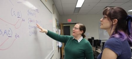 A professor writes equations on a whiteboard while a student looks on.