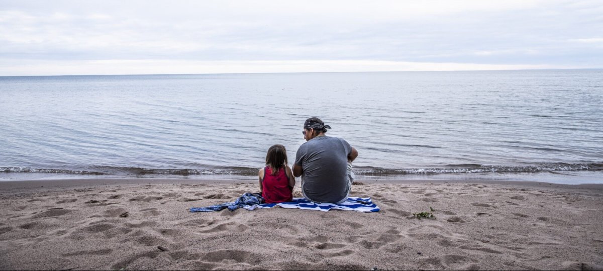 An adult and child sitting on a towel in the sand in front of the lake.