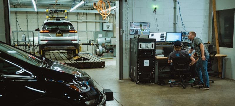 Two men look at computer screens in an automobile garage with two cars. 