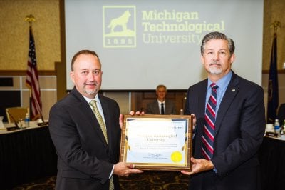 Two men in suits hold a framed award in a ballroom.