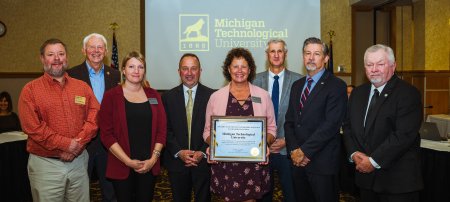 Eight staff members, board members and government officials stand in a ballroom smiling at the camera following the presentation of a framed award, held by a woman staff member in the center of the picture.