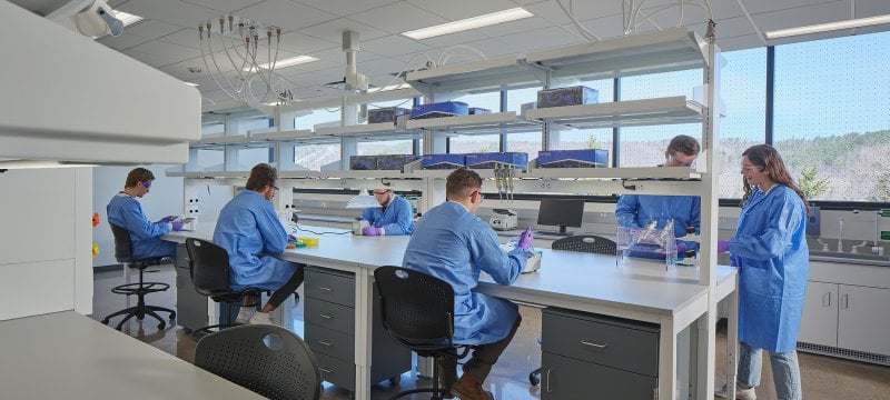 Six students in blue lab coats, purple gloves, and eye protection handle various medical equipment at a lab table. 