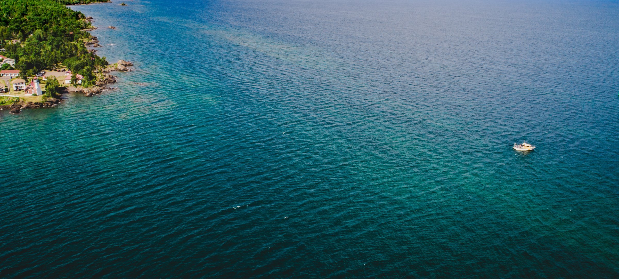 Drone aerial view of blue Lake Superior water on sunny day with small strip of green trees and shoreline on left and research boat in bottom right corner. 