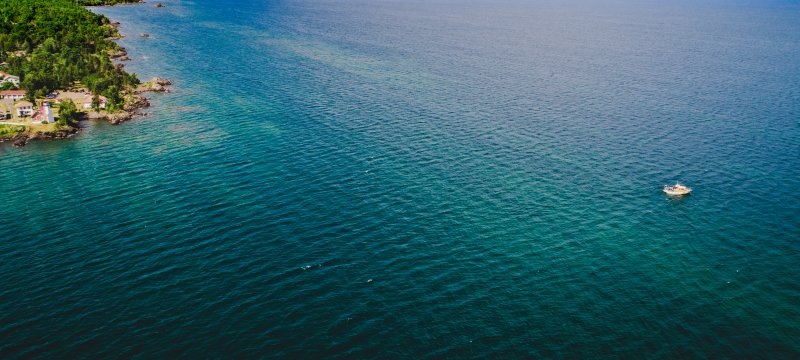 Drone aerial view of blue Lake Superior water on sunny day with small strip of green trees and shoreline on left and research boat in bottom right corner.