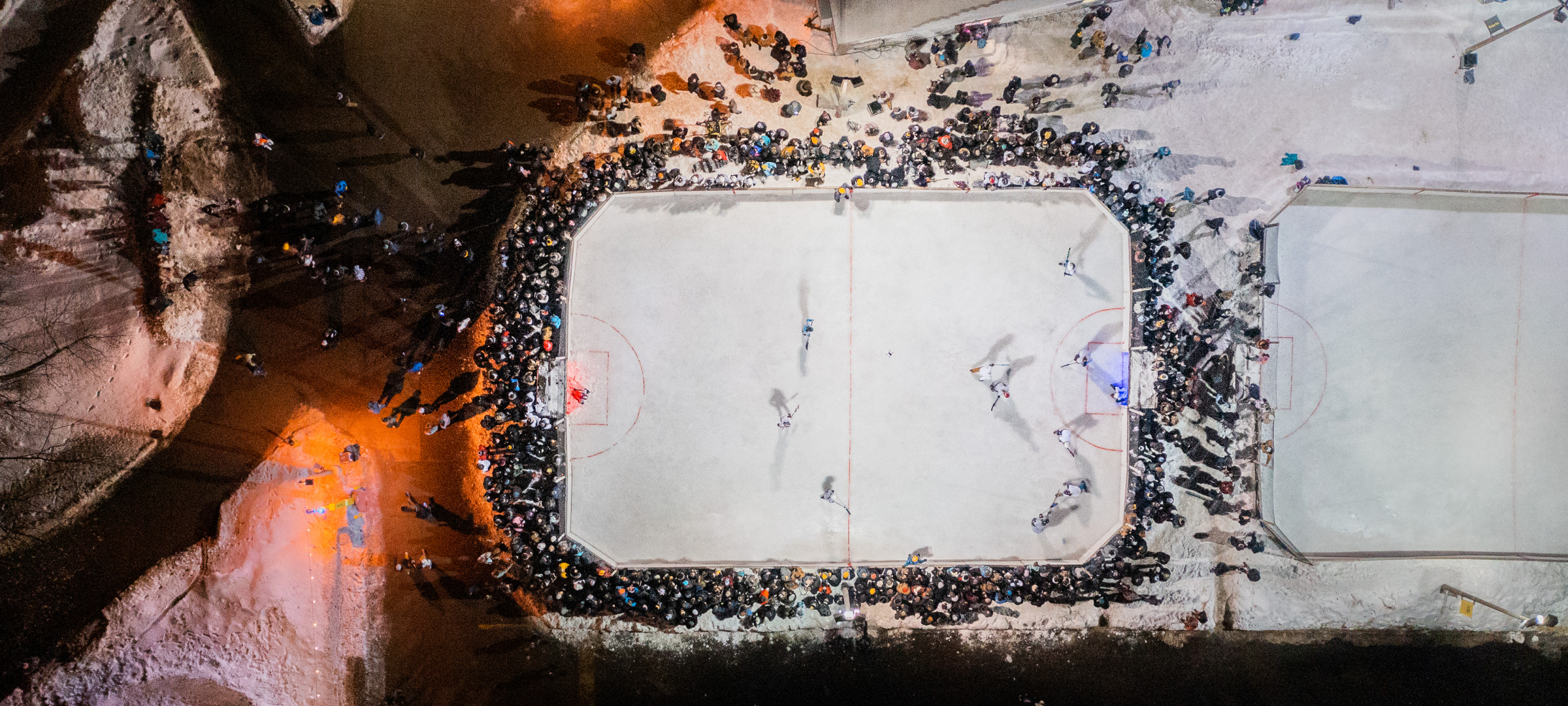 Aerial shot at night of dozens of students crowded around broomball rink in winter watching a game.