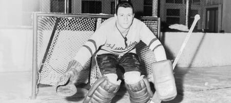 Black and white photo of dark-haired hockey goalie in Huskies sweater crouching with legs and arms out in front of a hockey goal, looking at the camera.