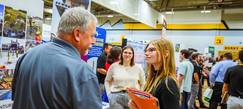 Gray-haired man in blue polo shirt speaks to female student wearing eyeglasses and smiling while holding an orange folder with resumes and employer information in a gymnasium with other students in business attire in background.   