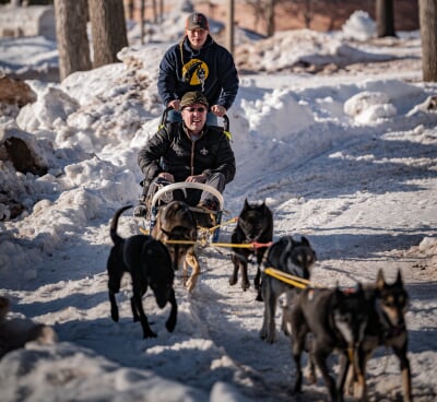 Brynn Santi on the back of a sled with a passenger being pulled by Mushing Club dogs.