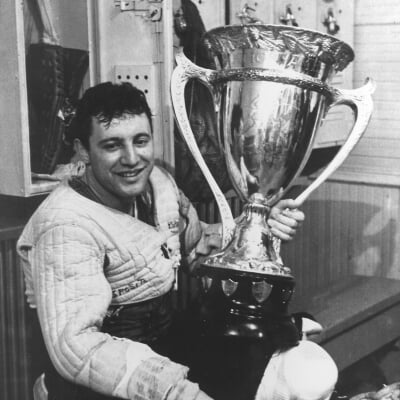 Black and white photo of MTU goalie Tony Esposito sits in locker room smiling at camera with large silver trophy in his lap.