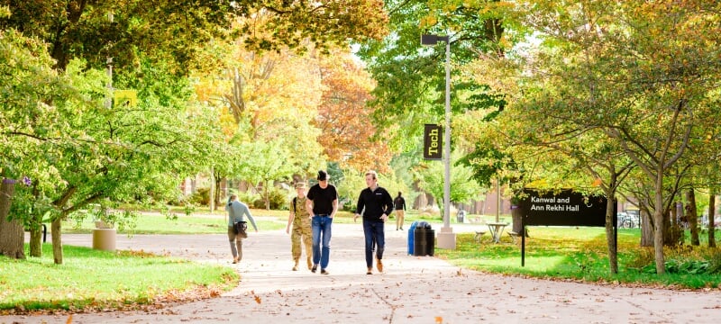 Five students walk and talk on campus sidewalk in the center of the frame with leaves in early fall colors overhead and in the background.