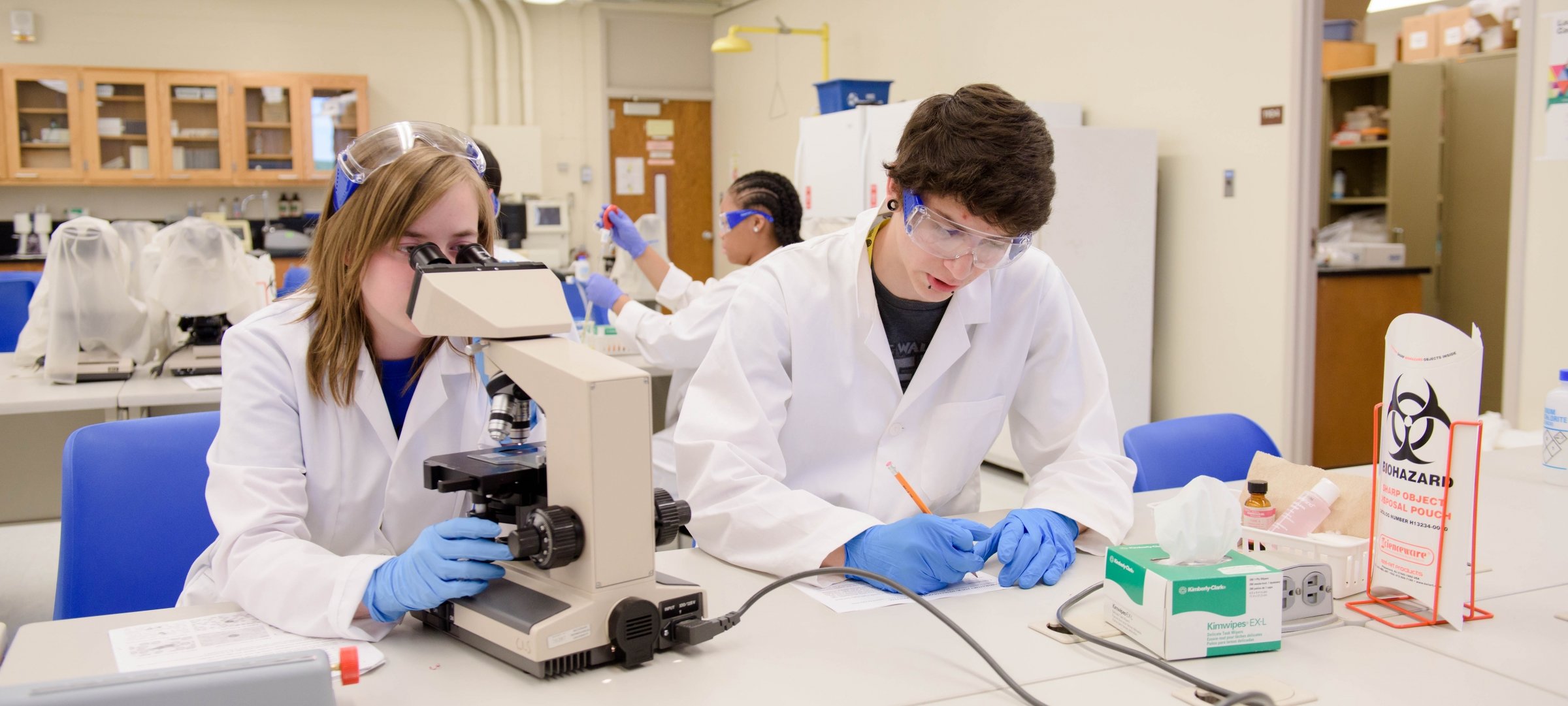 Two students in a lab sitting at a table, one taking notes, one looking through a microscope.