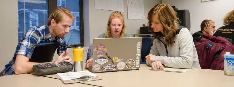 Three students interacting with a laptop in a classroom