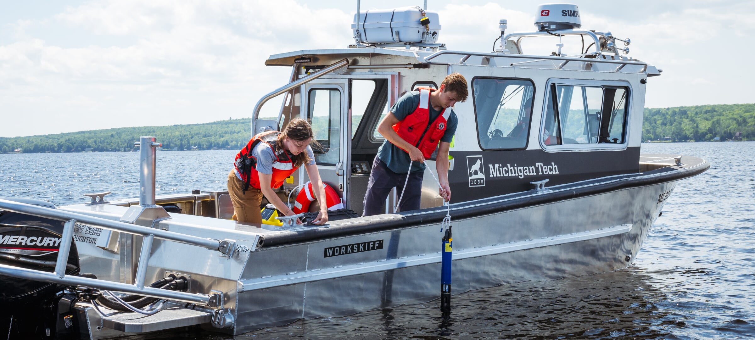 Two researchers with life vests lowering an instrument into the water from an MTU research vessel.