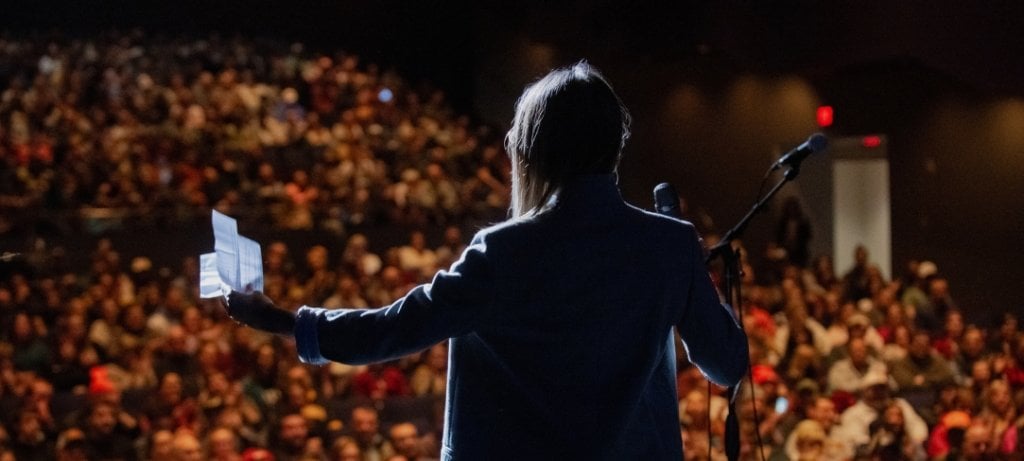 The back of a figure gesturing while giving a speech to a full audience.