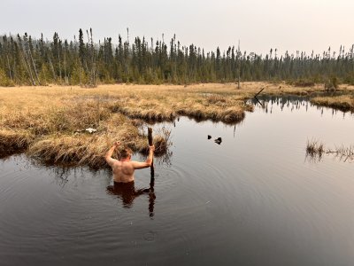 A man with a staff waist-deep in water with his back toward the camera and arms up wades toward bones in a marshy bog on a mat of grass in a clearing.