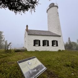 A white lighthouse on a small grassy hill with foggy waters behind it and a sign about the lighthouse in the foreground.