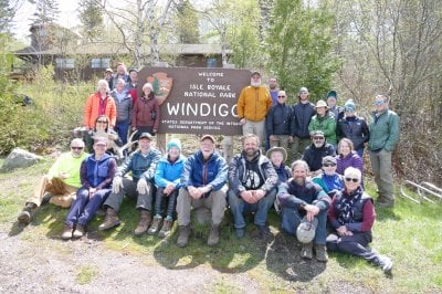 27 people gathered around a sign reading Isle Royale Windigo Welcome to Isle Royale National Park, States Department of the Interior, National Park Service, with one holding moose antlers.