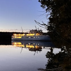 The lights of the Isle Royale Ranger III passenger ferry are reflected in the water in the darkening evening.