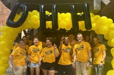 Ruby in a group of students wearing bright yellow It's Mac Night tee shirts under a ring of yellow balloons and black balloons spelling DHH.