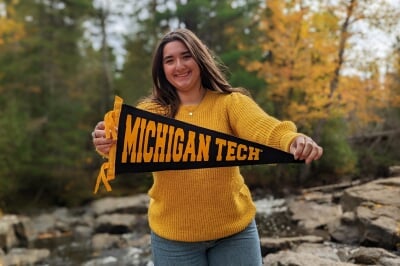 Ruby standing outside by a rocky brook holding a black Michigan Tech pennant.