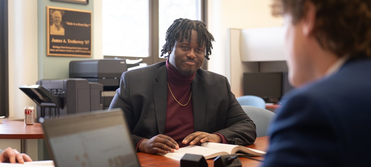 Elijah Joseph and other members of the Applied Portfolio Management Program conduct buy reports in the College of Business Trading Room. Elijah Joseph sitting at a table with a book. Other people are visible to the sides.