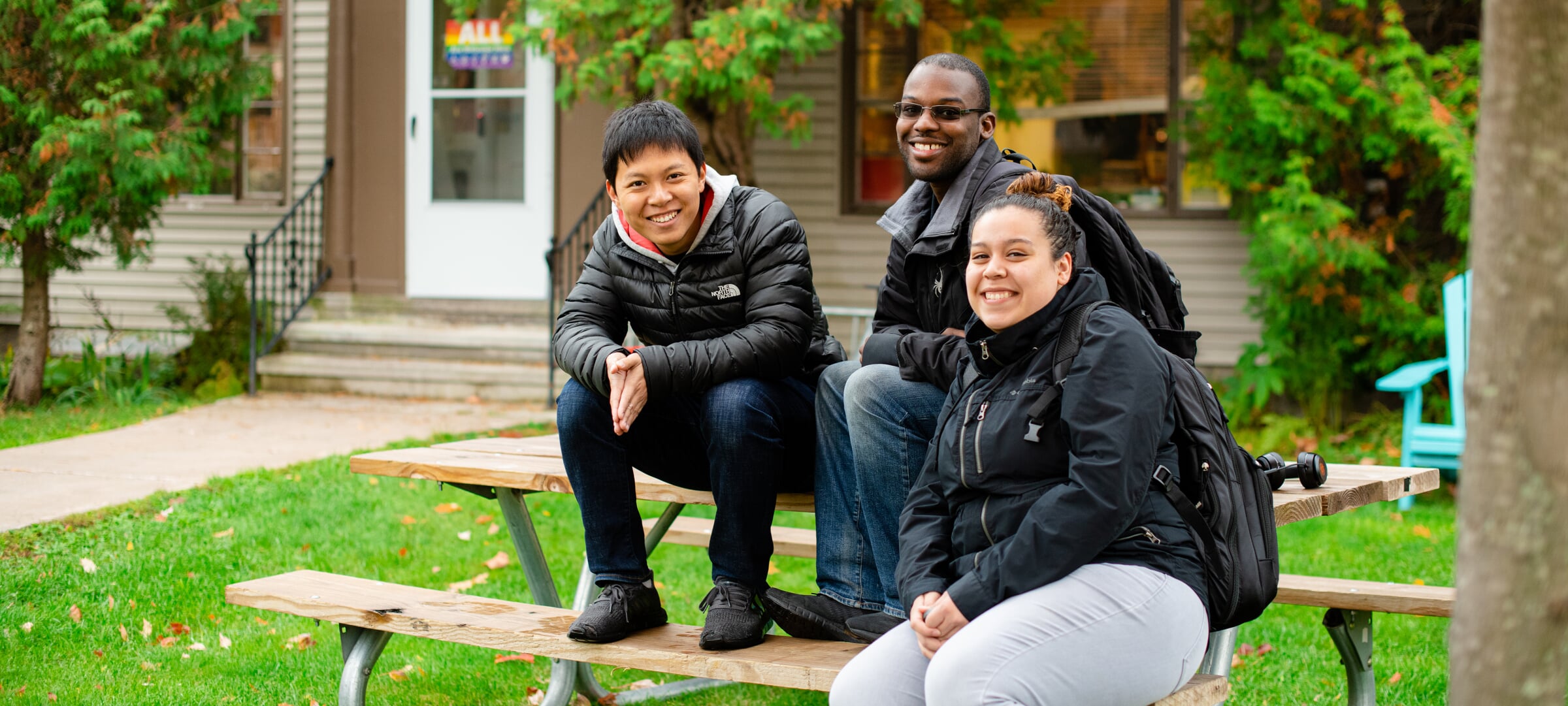 Three students sitting outside on a picnic table in front of the Hamar House at Michigan Tech.