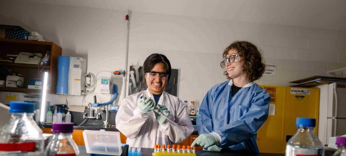 Two students stand in a chemistry lab with test tubes and a pipette.