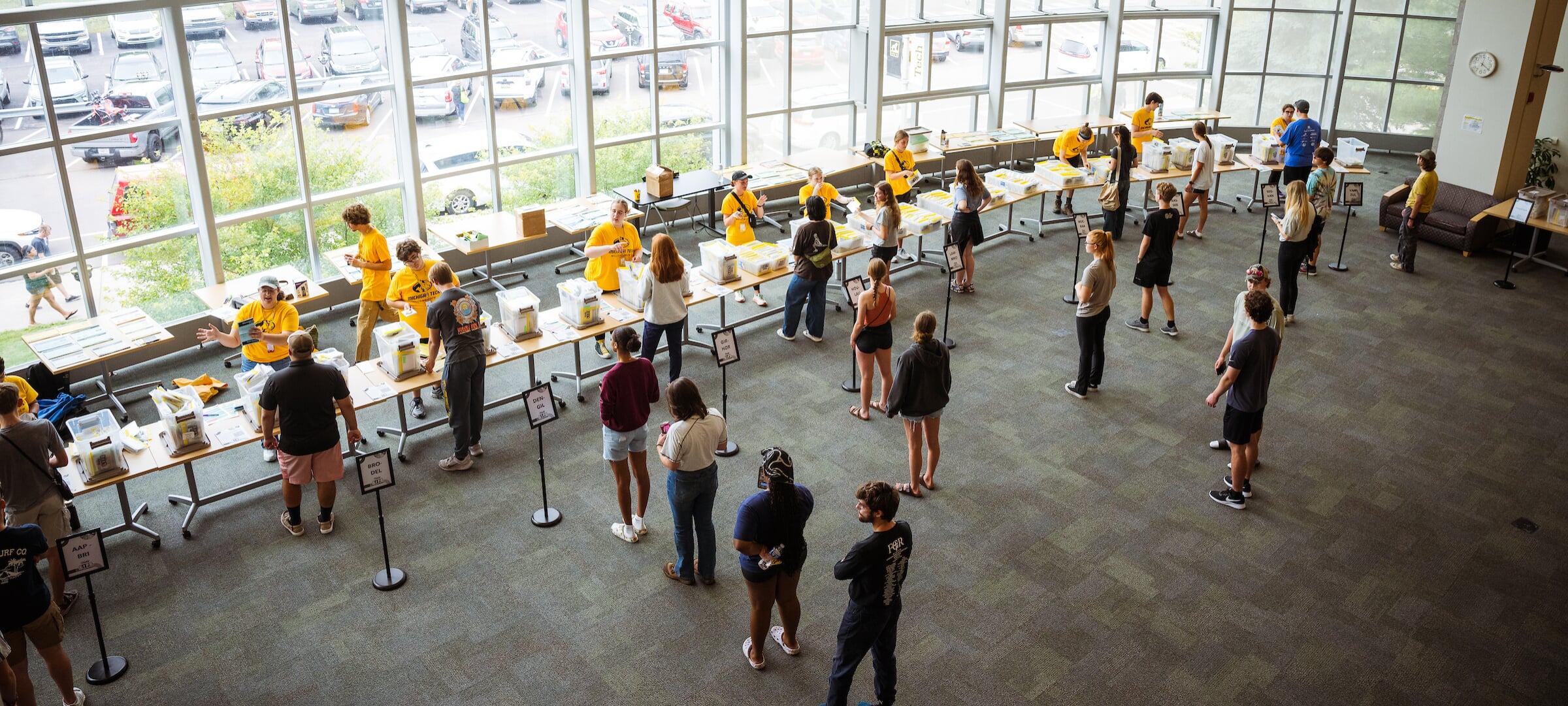 People in bright yellow shirts staff stand at tables in a modern glass-walled building lobby while new students and families line up to check in.