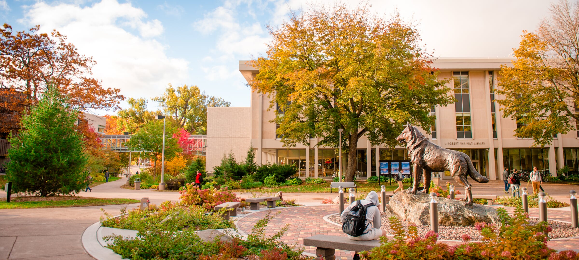 A person sits on a bench in front of Michigan Tech's husky statue and campus library.