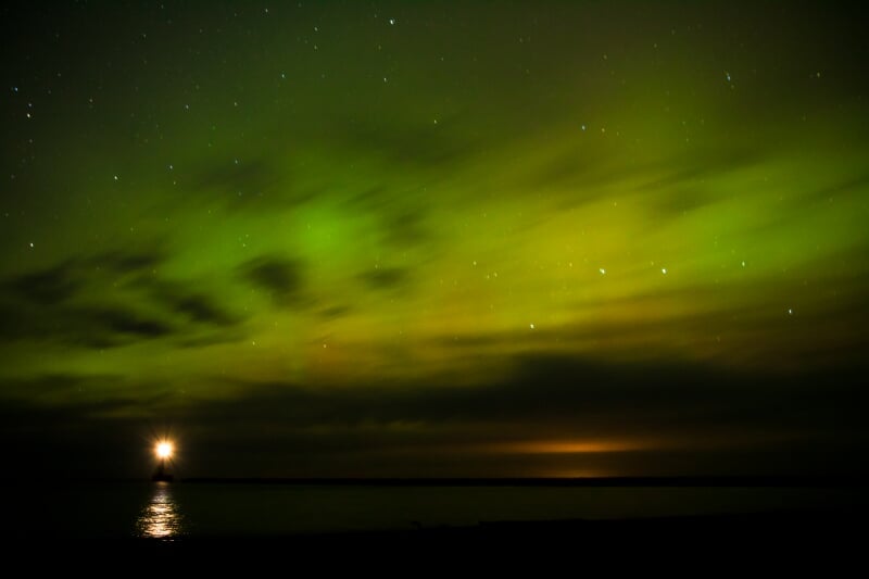 Northern Lights (Aurora Borealis) streaking green and yellow above a lake and lighthouse.