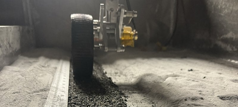 Straight-on view of a lunar rover wheel going over asphalt in the middle of sand.