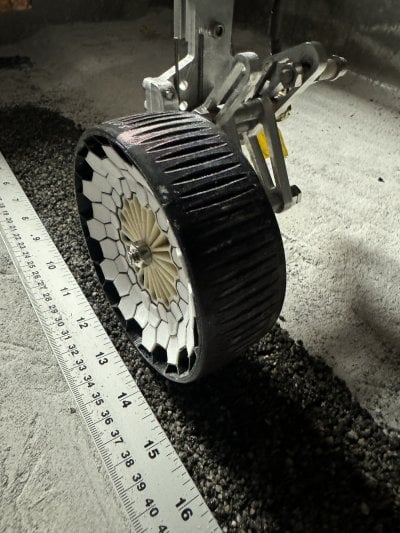 The wheel of the lunar rover on asphalt in the midst of sand next to a ruler.