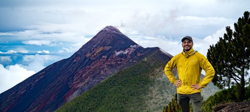 Man standing hands on hip in a yellow jacket, smiling in front of Peak of Volcan de Fuego, a volcano, in the background.