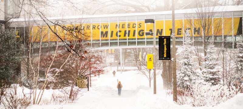 In winter, snow covers the ground under the raised, enclosed walkway between Van Pelt and Opie Library and Rekhi Hall, with signage reading “Tomorrow Needs Michigan Tech.”